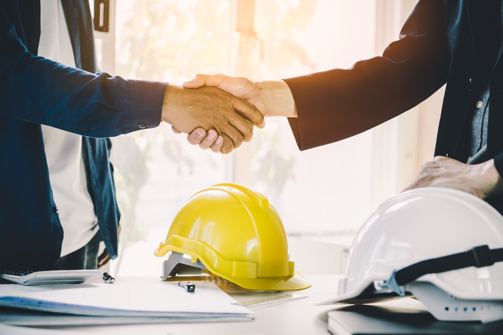 Hands shaking in a professional setting, with a yellow hard hat and a white hard hat on a table nearby, symbolizing a construction agreement.