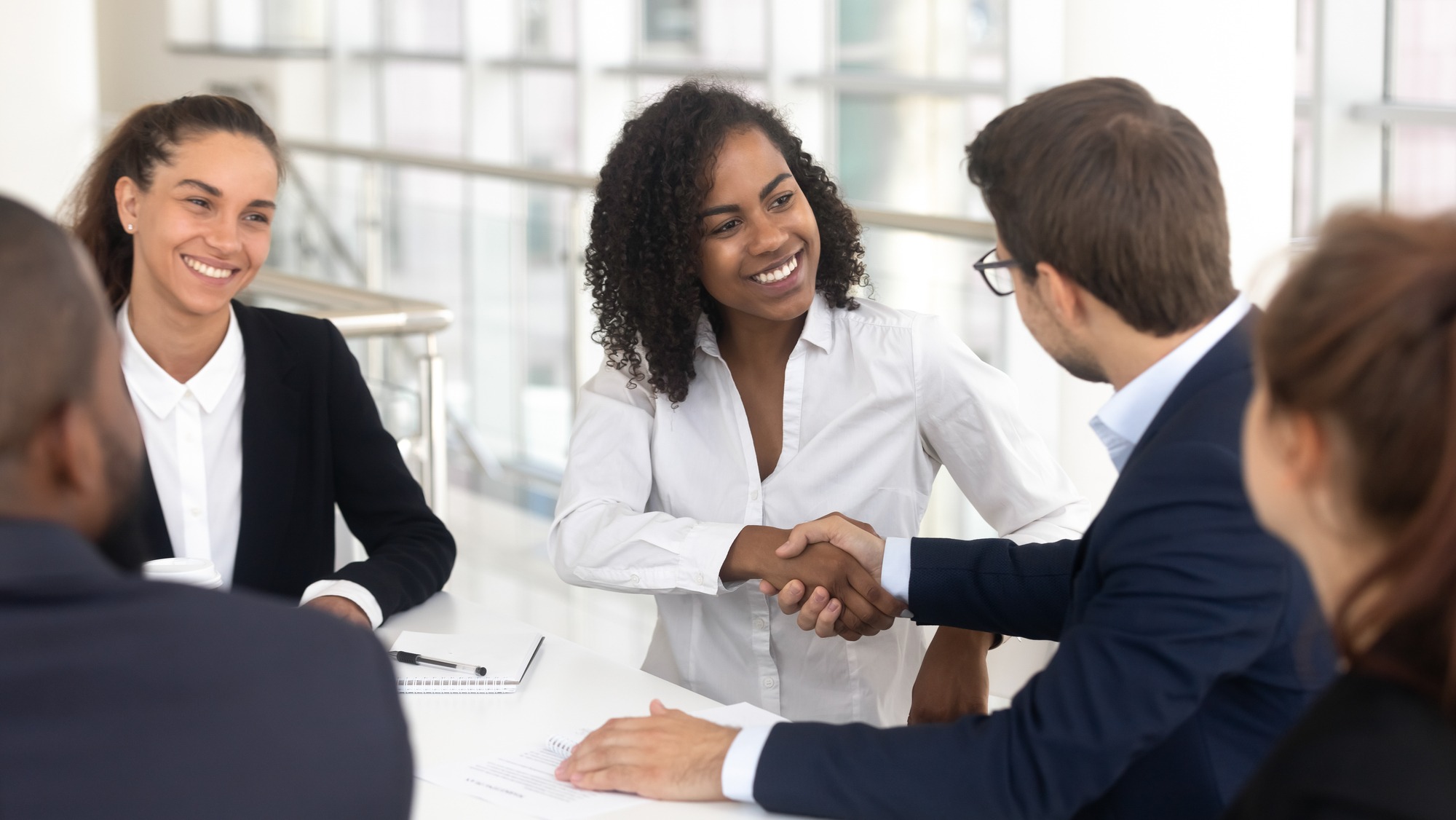 A business meeting showing a woman smiling and shaking hands with a man, surrounded by colleagues, highlighting collaboration and professionalism.