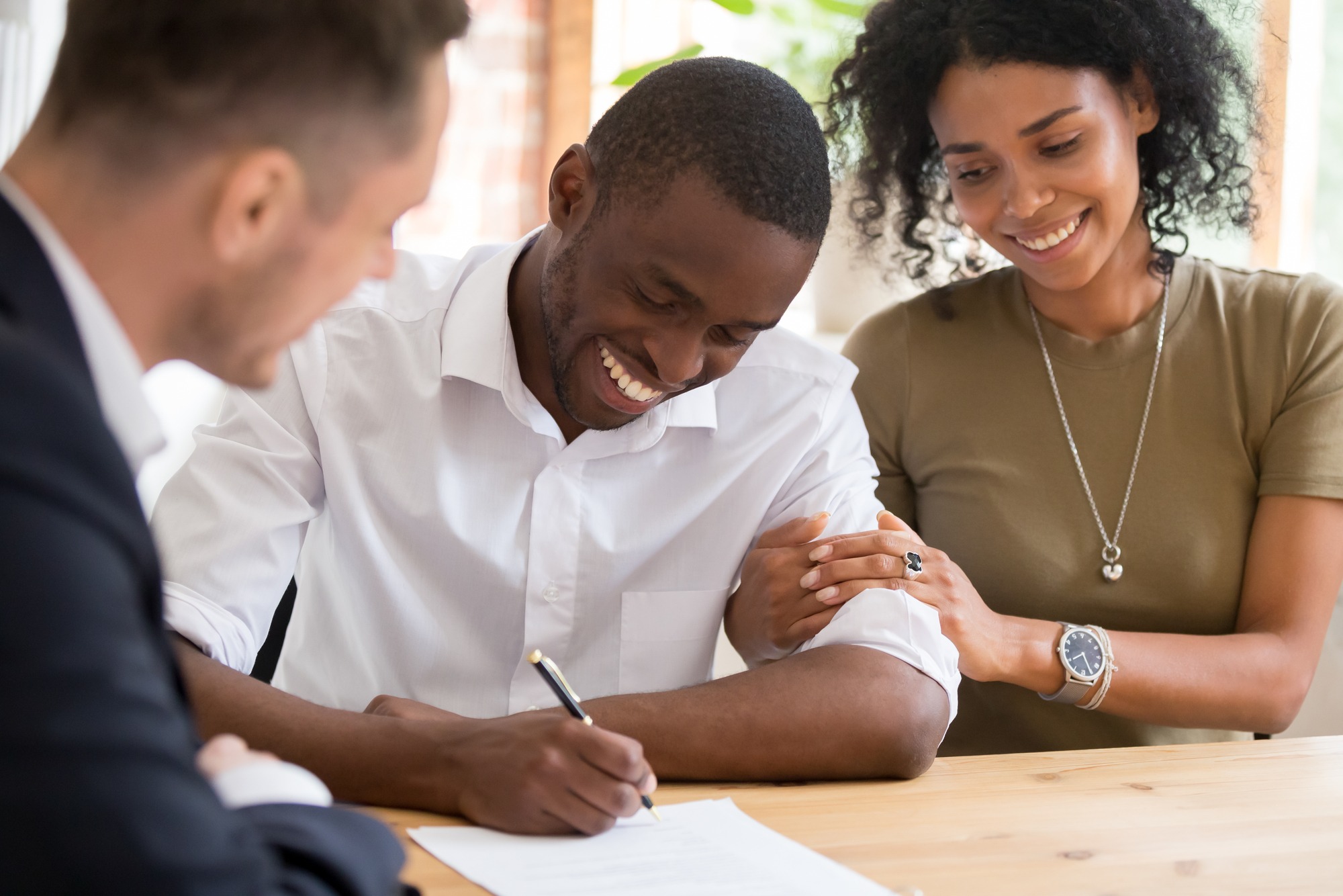 A man joyfully signs a document while a woman shares a supportive moment beside him. Another person observes, fostering a collaborative atmosphere.