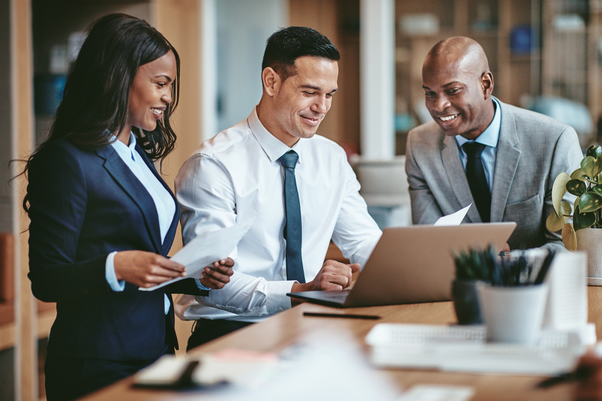 Three professionals collaborate at a laptop in a modern office, discussing documents and sharing insights, highlighting teamwork and productivity.