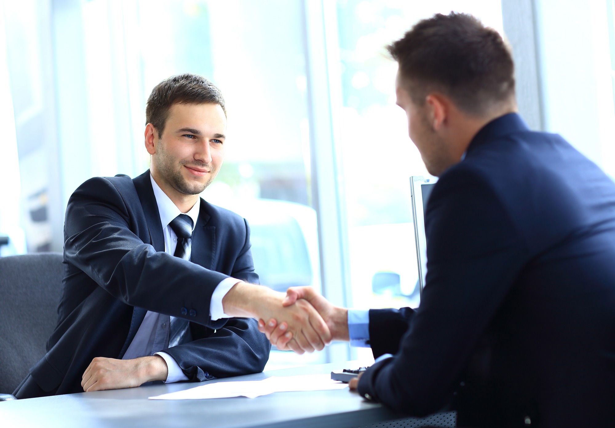 Two men in business suits shake hands across a desk in a modern office setting, symbolizing a successful agreement or partnership.
